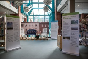 Ausstellungsansicht mit Medienstationen, Text-Bild-Tafeln und verschiedene Objekte. Darunter eine Sitzgruppe aus alten Gartenmöbeln.