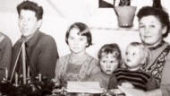 Zwei Erwachsene und drei Kinder sitzen an einem Tisch. Vor ihnen stehen ein Adventskranz sowie zwei Teller, die mit gebackenen Keksen gefüllt sind.