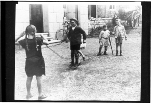 Fünf Kinder vor einem Haus auf der Dorfstraße. Zwei von ihnen halten ein Seil, über das zwei andere Kinder springen.