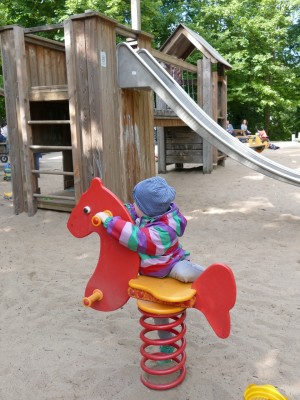 Ein Kind mit bunt gestreifter Jacke und Mütze klettert auf ein rotes Feder-Pony im Sand stehend auf einem Spielplatz. Im Hintergrund ist ein Klettergerüst mit Rutsche und Hängebrücke zu sehen.
